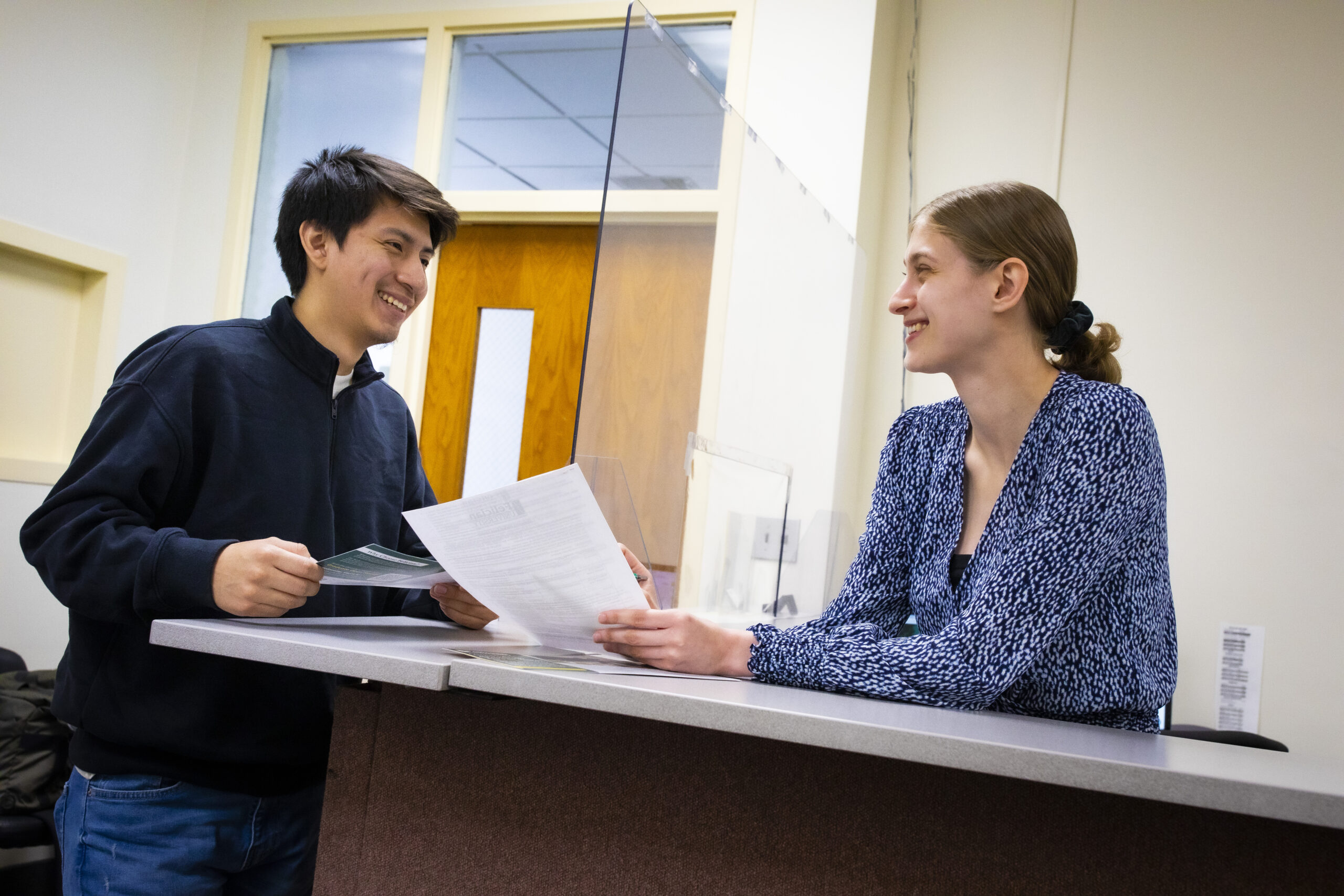 Woman leans over desk, holding paper, smiling to man while pointing something out on the paper